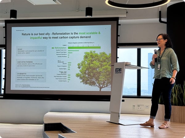 An impact investor stands on stage with a large presentation screen next to her, speaking to other investors